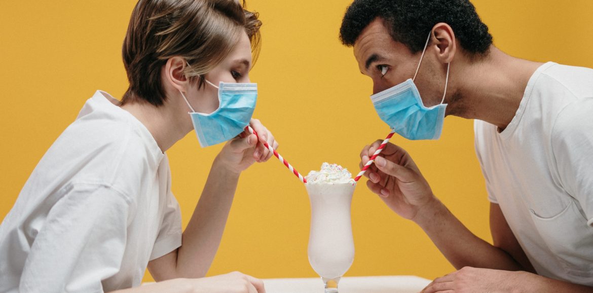 Une femme et un homme boivent un milkshake dans le même verre avec leur paille, tout en portant un masque anti-Covid.