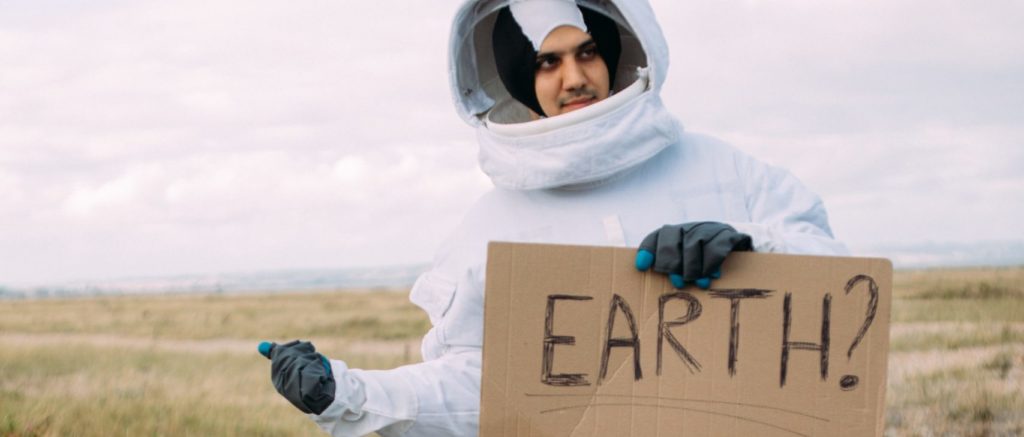 Un homme habillé en cosmonaute fait du stop en tenant une pancarte en carton qui indique "Earth ?" 