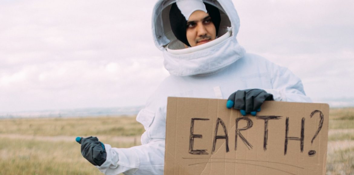 Un homme habillé en cosmonaute fait du stop en tenant une pancarte en carton qui indique "Earth ?"
