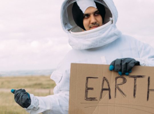 Un homme habillé en cosmonaute fait du stop en tenant une pancarte en carton qui indique "Earth ?"