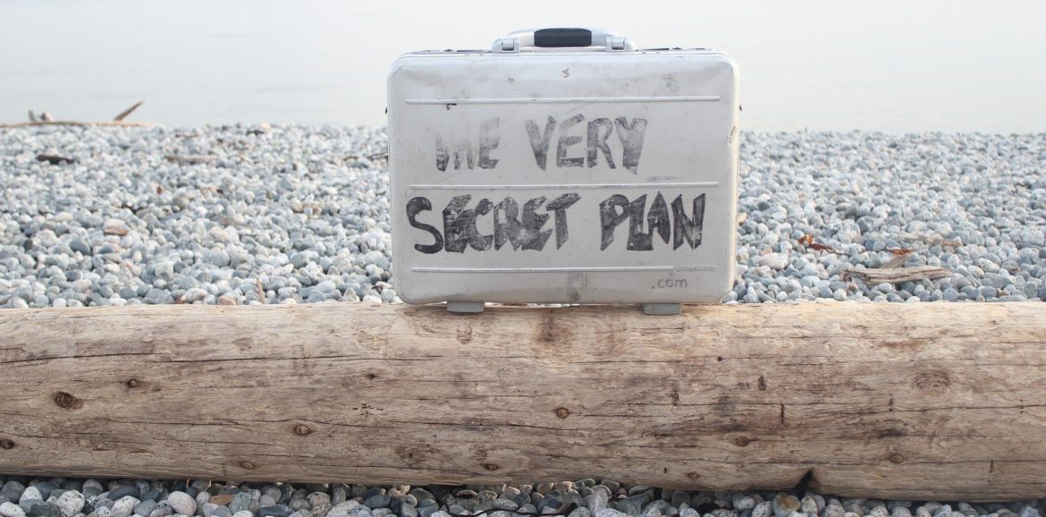 Valise posée sur une plage de galets portant l'inscription "The very secret plan"