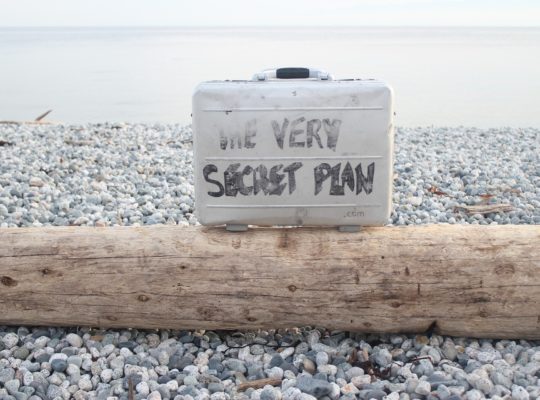 Valise posée sur une plage de galets portant l'inscription "The very secret plan"